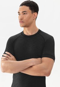 Zwart sportshirt met korte mouwen en een ronde hals, gladde textuur en contrasterende raglanmouwen. Model poseert met gekruiste armen.