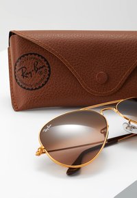 Ochelari de soare Ray-Ban cu lentile ovale de culoare maro, ramă metalică aurie și suporturi pentru nas transparente. Însoțiți de un etui texturat de culoare marron cu un logo.