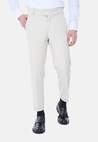 Pantaloni beige con un fit su misura, chiusura con zip e bottone. Presentano tasche laterali e una texture liscia, abbinati a scarpe nere.