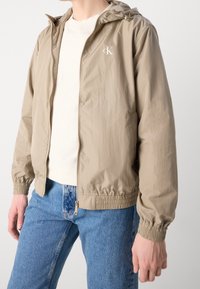 Béžová lehká bunda s kapucí, elastickými manžetami a lemem, zapínáním na zip a bílým logem na hrudi. Nošena přes světle barevnou košili.