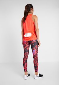 Femme debout, vue de dos, portant un haut sans manches orange vif et un legging à motifs rouges avec des chaussures de sport noires et blanches.