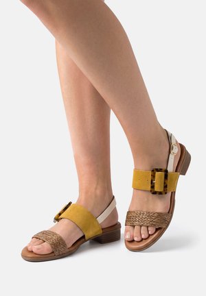 Pieds portant des sandales à bouts ouverts avec une lanière avant tissée, une lanière médiane en daim jaune moutarde avec une boucle écaille, et une lanière de cheville blanche.
