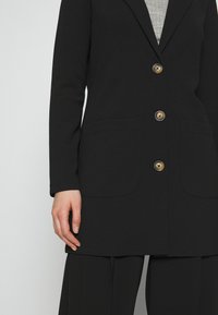 Černý blazer s přiléhavým střihem, který má klopu, tři zlaté knoflíky a dvě přední kapsy. Hladká tkanina a minimalistický design.
