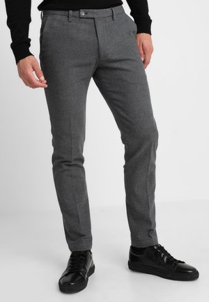 Man draagt slanke grijze pantalon en zwarte sneakers, staand met ontspannen handen langs het lichaam tegen een effen witte achtergrond.