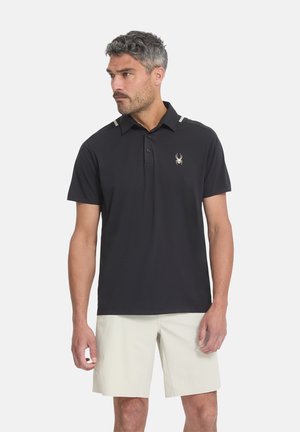 Homme aux cheveux gris portant un polo noir à manches courtes avec un logo araignée et un short beige, debout devant un fond uni.