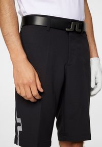 Svarta golfshorts i ett glatt tyg, med ett vitt randmönster på sidan, i kombination med ett svart läderbälte och handskar.