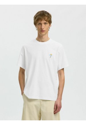 Młody mężczyzna o jasnobrązowych włosach, ubrany w biały T-shirt z małym motywem palmy i słońca, beżowe szorty, stojący na białym tle.