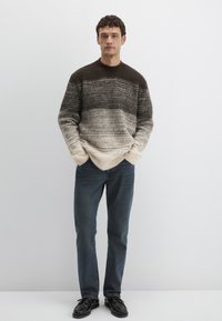 Gestrickter Pullover mit Verlaufsmuster in Dunkelbraun, Beige und Creme, kombiniert mit dunkelblauen Jeans und schwarzen Schuhen.