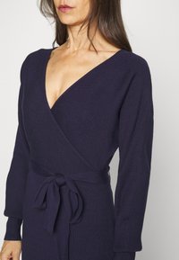 Femme portant une robe portefeuille côtelée bleu marine à manches longues avec une ceinture nouée à la taille, montrée du cou aux hanches sur un fond uni.
