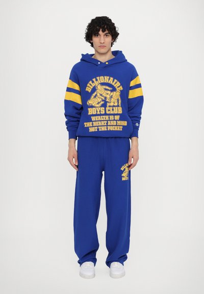 Blaue Baumwollkapuzenjacke mit gelben Streifen und grafischem Druck, kombiniert mit blauen Sweatpants mit einem Logo. Weiße Sneakers an den Füßen.