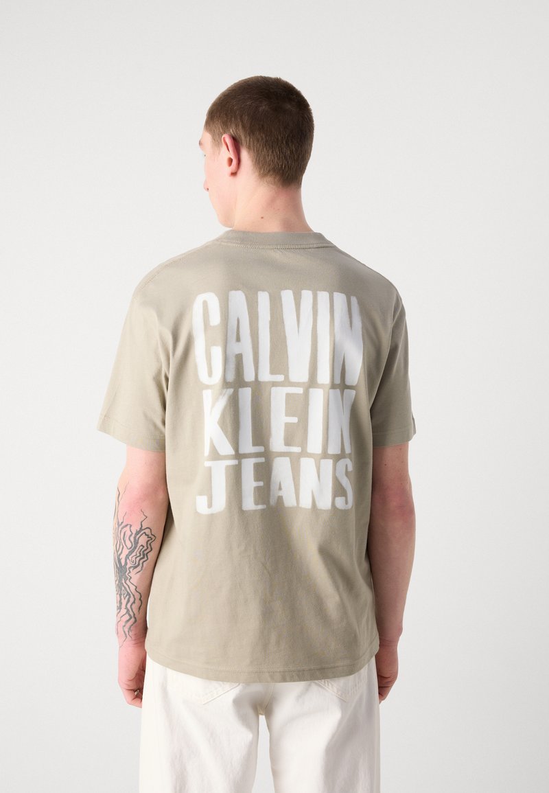 Siva pamučna majica s velikim bijelim natpisom "CALVIN KLEIN JEANS" na leđima. Kratki rukavi i okrugli izrez. Glatka tekstura.