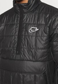 Zwarte pufferjack met een hoge kraag en een halve rits. Kenmerkt zich door een gewatteerde textuur en een wit Nike-logo op de linkerborst.