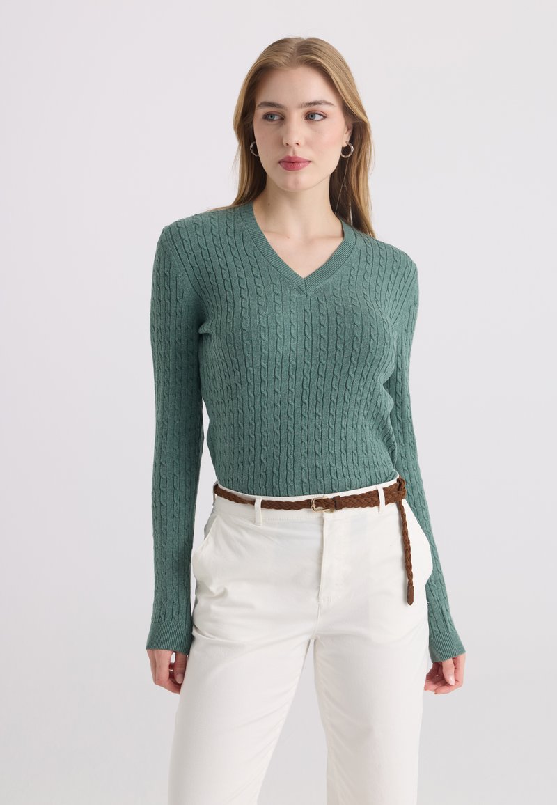 Femme portant un pull vert à col en V en maille torsadée, pantalon blanc et ceinture tressée marron, debout devant un fond clair uni.