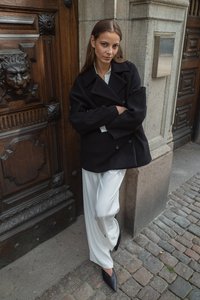 Musta kaksirivinen takki valkoisen paidan päällä, yhdistettynä vaaleisiin leveälahkeisiin housuihin ja mustiin kärkitossuihin. Kivinen tausta.