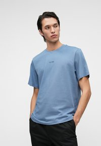 Kék pamut póló rövid ujjakkal és kerek nyakkivágással. A mellrészén egy kis fekete "Calvin Klein" logó található. Simás textúra.