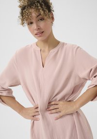 Rochie lungă, roz deschis, cu mâneci lungi și decolteu în V, dintr-un material texturat, având pliuri delicate și buzunare laterale, purtată cu mâinile pe șolduri.