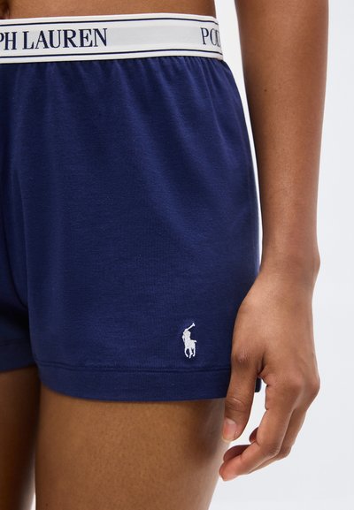 Σκούρα μπλε μποξερ με λευκή ελαστική ζώνη Polo Ralph Lauren στη μέση ενός ατόμου και το δεξί χέρι στο πλάι.