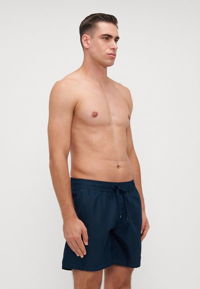 Pantaloni scurți de înot navy cu bandă elastică și șnur, având o textură netedă și lungime peste genunchi, modelul prezentat stând în picioare.