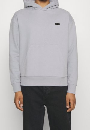 Mężczyzna ubrany w jasnoszarą bluzę z kapturem Calvin Klein z przednią kieszenią i czarne spodnie, pokazany od ramion do bioder.