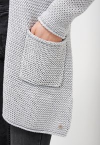 Světle šedý pletený cardigan s velkou přední kapsou a žebrovanými manžetami, s hladkou texturou a jemným značkováním na lemu.