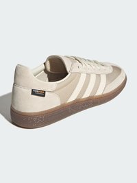 Ljusbeige sneaker med mesh- och mockamaterial, med tre vita ränder och en yttersula i gummiresår. Stor präglad logotyp på hälen.