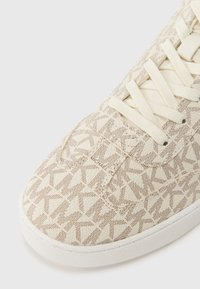 Μπεζ αθλητικό παπούτσι με υφή, χαρακτηριστικό σχέδιο με κορδόνια και λευκή καουτσούκ σόλα. Λογότυπο τυπωμένο στο πλάι. Ομαλή επιφάνεια συνολικά.