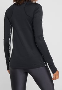 Frau trägt ein schwarzes, langärmliges Sportoberteil mit dem Text "Nike Pro" auf den Ärmeln, kombiniert mit glänzenden schwarzen Leggings, von hinten gezeigt.