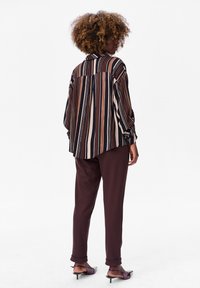 Striped oversized blouse in black, brown, and cream; paired with maroon pants. Slender heeled shoes; smooth fabric with relaxed fit and cuffs.