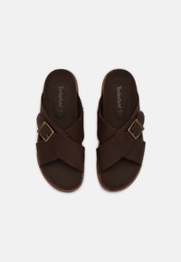 Sandales en cuir marron avec un design de brides croisées, dotées d'une boucle en métal et d'un semelle intérieure texturée douce. Aspect minimaliste et décontracté.