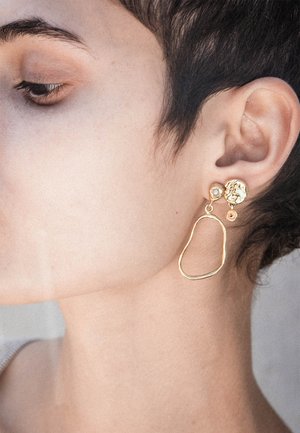 Guldøreringe med en struktureret rund stud, med en hængende uregelmæssig formet ring. Designet kombinerer glatte og bølgede elementer.