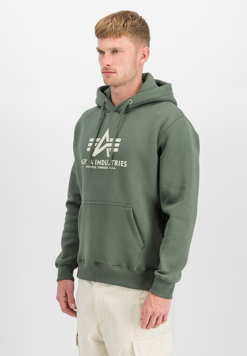 Jeune homme portant un sweat à capuche vert Alpha Industries et un pantalon cargo beige, se tenant devant un fond blanc uni.