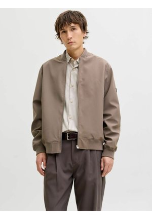 Ung mann iført en taupe bomberjakke over en beige stripete skjorte med knapper og mørkebrune bukser, stående mot en enkel, lys bakgrunn.