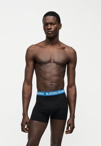 Άνδρας μοντέλο στέκεται σε απλό φόντο φορώντας μαύρα μποξέρ με μπλε ζώνη μέσης "BJÖRN BORG", τα χέρια χαλαρά στο πλάι.