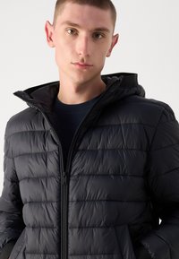 Sort puffjakke med lynlås foran, høj krave og vandret quiltet design. Glat tekstur med tætsiddende manchetter og hætte.