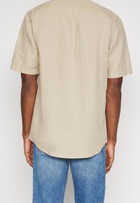 Persona che indossa una camicia beige a maniche corte e jeans blu, vista da dietro su uno sfondo chiaro e uniforme.