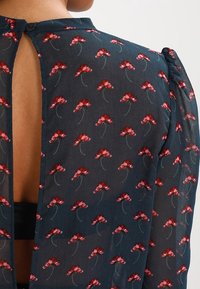 Blouse fleurie en tissu bleu foncé transparent avec un motif répétitif de fleurs rouges, fermeture par boutons au niveau du cou et dos ouvert.