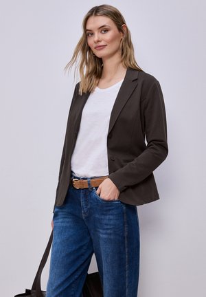 Femme portant un blazer noir, une chemise blanche, un jean bleu avec une ceinture marron, tenant la sangle d'un sac noir, debout devant un fond gris clair.