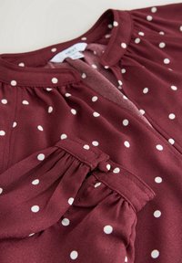 Blouse marron avec des pois blancs, col rond et détail froncé près du poignet boutonné.