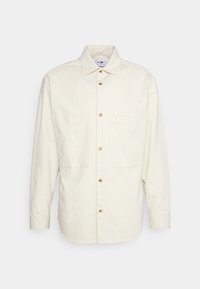 Chemise longue à manches longues crème clair avec un col pointu, deux poches poitrine et des boutons dorés. Fabriquée en tissu texturé.
