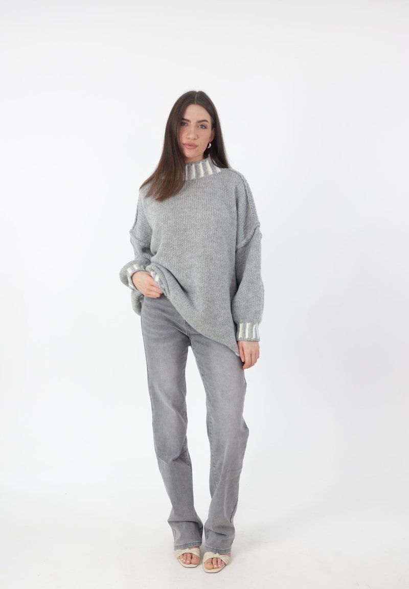 Grijze gebreide trui met een hoge kraag en zilverkleurige accenten op de mouwen, gedragen met lichtgrijze flare broek. Glad oppervlak, ontspannen pasvorm.