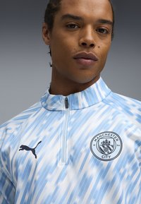 Helles blaues Trainingsoberteil mit Reißverschluss und diagonalen Streifen, mit einem schwarzen Puma-Logo und dem Wappen von Manchester City auf der linken Brust.