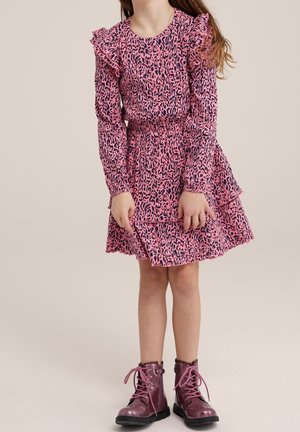 Fille portant une robe à imprimé léopard rose et noir avec des épaules volantées et des bottes à lacets roses brillantes, debout devant un fond uni.
