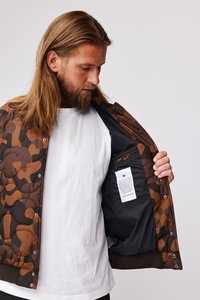 Brun camouflage quiltet jakke med trykknaplukning, foret indre med lommer og et plejetikett synligt indeni.
