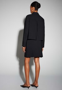 Zwarte getailleerde jas met plooi-detail, gecombineerd met een nauwsluitende zwarte rok. Gladde stoftextuur, minimalistisch ontwerp en puntige zwarte mules.
