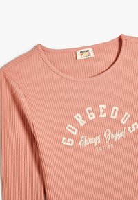 Roze geribbelde lange mouw shirt met een ronde halslijn. Bevat witte tekst met de tekst "GORGEOUS Always Joyful EST 92." Zachte stoftextuur.