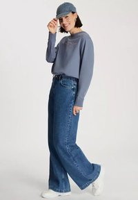 Jasnoniebieska bluza z nadrukowanym tekstem, zestawiona z szerokimi niebieskimi dżinsami i pasującą dżinsową czapką, uzupełniona białymi sneakersami.