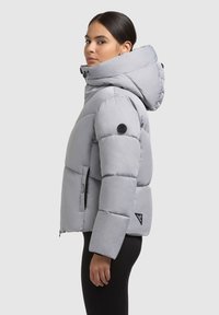 Giacca a vento grigia con colletto alto, cappuccio e polsini a coste. Presenta una patch con logo nero e tasche laterali con zip. Texture liscia e design leggero.