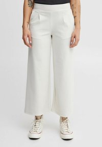 Pantaloni bianchi a gamba larga realizzati in tessuto morbido, con pieghe frontali e vita alta, abbinati a sneakers di colore chiaro.