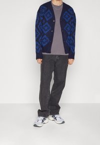 Marinblå kofta med geometriska blå mönster, knäppning framtill och ribbade ärmslut, kombinerad med mörkgrå byxor och texturerade sneakers.