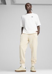 Hvid oversized t-shirt med sort logo, cremefarvede sweatpants og beige sneakers med grønne accenter. Glat tekstur, afslappet design.
